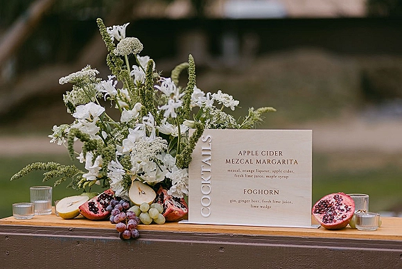 Wedding cocktail sign and cocktail menu sign wedding on a wood stand with white florals, fruit garnish, and votive candles on an outdoor lawn backdrop