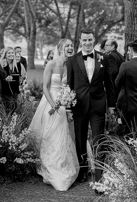 Wedding recessional as bride and groom walking hand in hand, bride laughing with a veil, bouquet, and guests cheering along a floral garden path