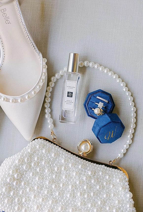 Bridal accessories flatlay with white pointed-toe shoe, pearl necklace and clutch, perfume bottle, and engagement rings in a blue velvet box on light fabric