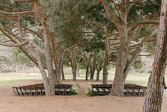 Outdoor ceremony setup with wood folding chairs and white floral aisle markers arranged in the round on a grassy pine tree grove lawn