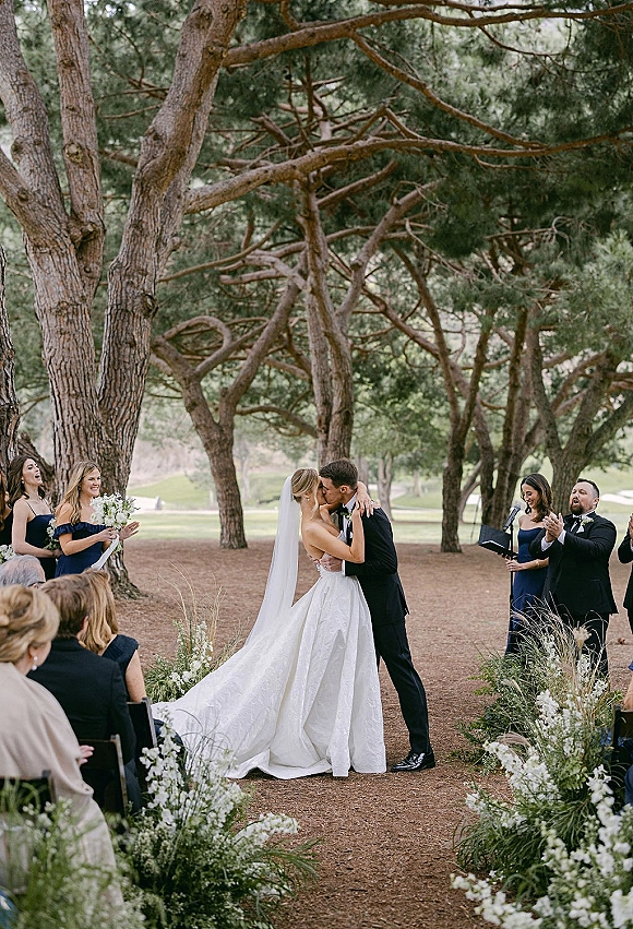 Ceremony kiss as bride in a long veil and groom in a black tuxedo kiss on a dirt aisle lined with florals in a tree grove ceremony