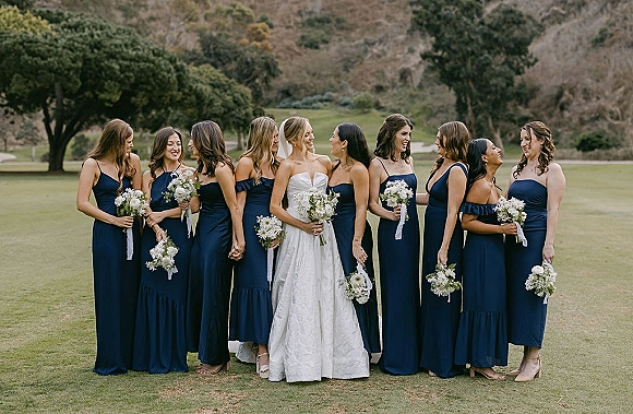 Bridesmaids group photo with bride with bridesmaids in navy dresses, holding white bouquets, standing on a grassy lawn with hills and trees behind
