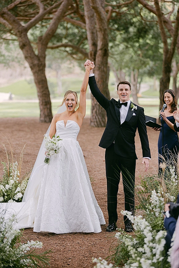 Ceremony moment as newlyweds raise hands and walk down a dirt aisle, bride in veil with bouquet beside groom in tuxedo under trees