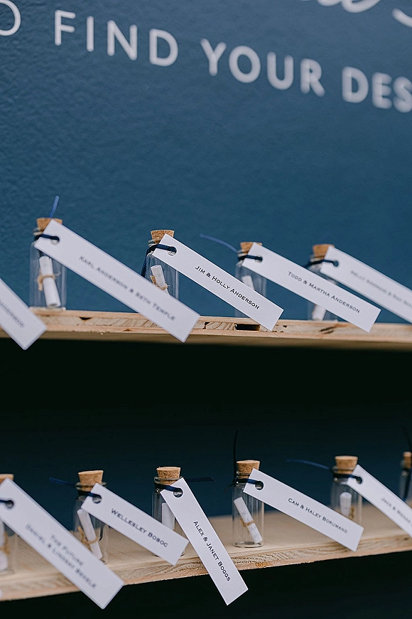 Wedding place cards in corked glass bottles with mini scrolls and string on wooden shelves, set against a chalkboard sign backdrop