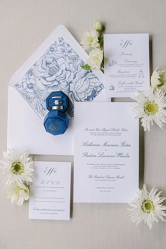 Invitation suite with blue and white wedding invitations, floral envelope liner, embossed cards, blue velvet ring box and rings on neutral fabric backdrop