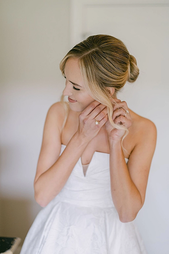 Bride portrait during bridal getting ready, looking down while adjusting an earring, wearing a strapless gown and pearl ring by a neutral wall