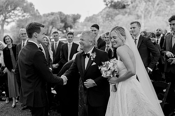 Ceremony moment as bride in veil holds bouquet, laughing while groom in tuxedo greets her father along a tree-lined outdoor aisle