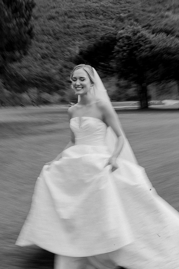 Bridal portrait in black and white of a bride twirling in a strapless gown with flowing veil and motion blur on a tree-lined lawn