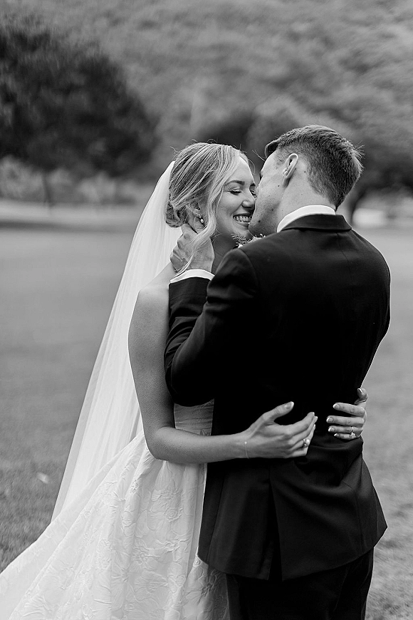 Wedding kiss portrait of bride and groom kissing as he holds her face, veil flowing over lace dress on a lawn with trees and hillside