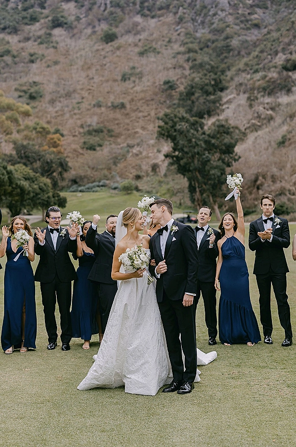 Wedding party photo of bride and groom kissing as the bridal party cheers, bridesmaids in navy dresses on a mountain hillside lawn