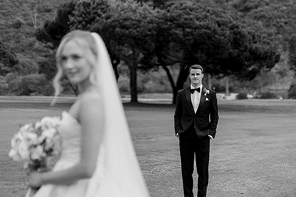 Wedding first look as the groom waiting first look in a tux watches the bride approach with long veil and white bouquet on a tree-lined lawn