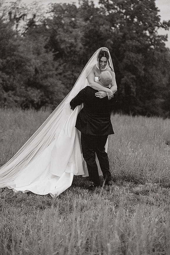 Couple portrait of groom lifting the bride as they embrace, her long cathedral veil draping over a strapless gown in a meadow with trees
