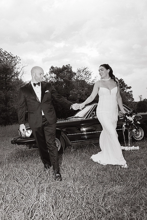 Couple portrait of bride and groom holding hands beside a classic car, her strapless dress and bouquet in a grassy field with trees