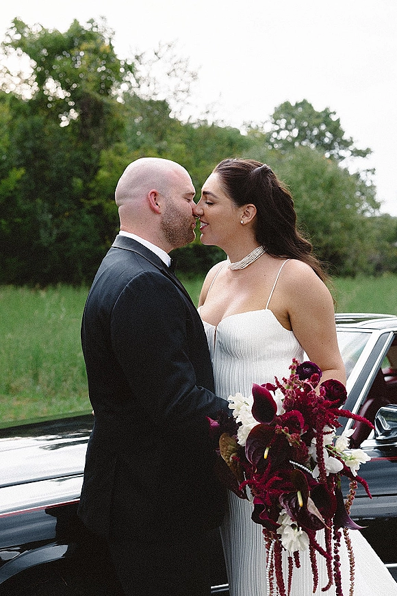 Wedding couple portrait of bride and groom nose kiss beside a vintage car, bride holding cascading burgundy and white calla lily bouquet outdoors