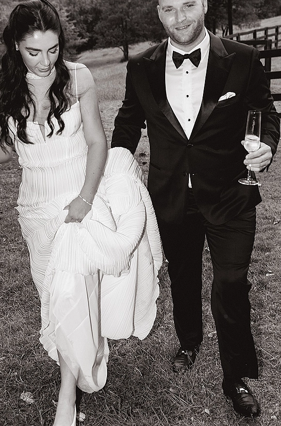 Wedding couple portrait in a black and white wedding photo, bride lifting her strapless gown beside groom with champagne flute on a lawn by trees and a wooden fence