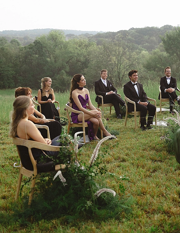 Wedding ceremony guests seated on wood chairs along a greenery aisle with white ribbon, in a grassy field with trees and hills