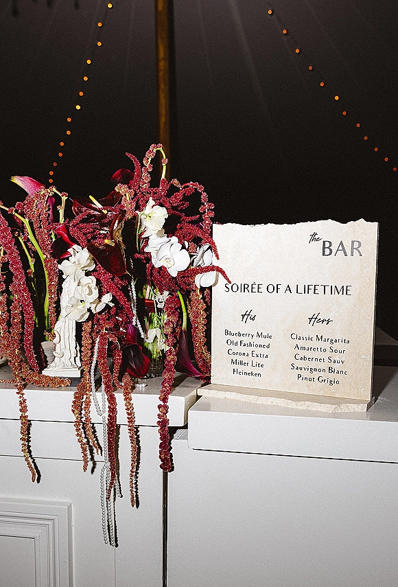 Wedding bar sign with wedding bar menu on a white counter, framed by calla lilies, orchids, hanging amaranthus and pearl accents under string lights