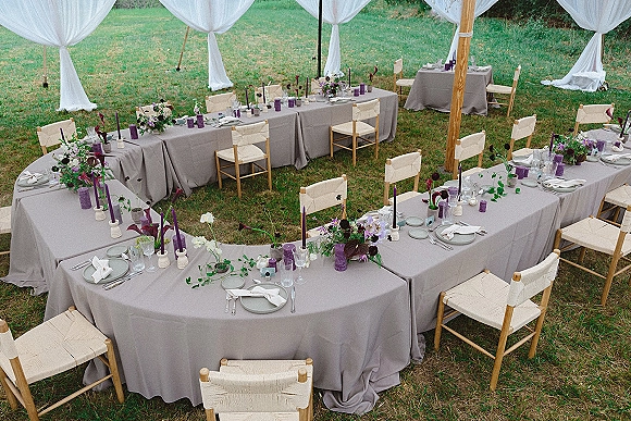 Reception tablescape with gray linens, purple taper candles and floral greenery garland on long tables under a white draped tent on a lawn