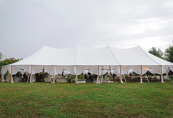 Wedding tent reception with a sailcloth wedding tent, draped curtains, string lights, banquet tables, bar and lounge seating on grass field