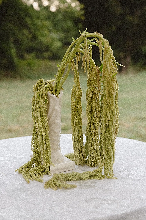 Floral centerpiece with hanging greenery in a white ceramic vase on a round table with white cloth, set on an outdoor lawn with trees