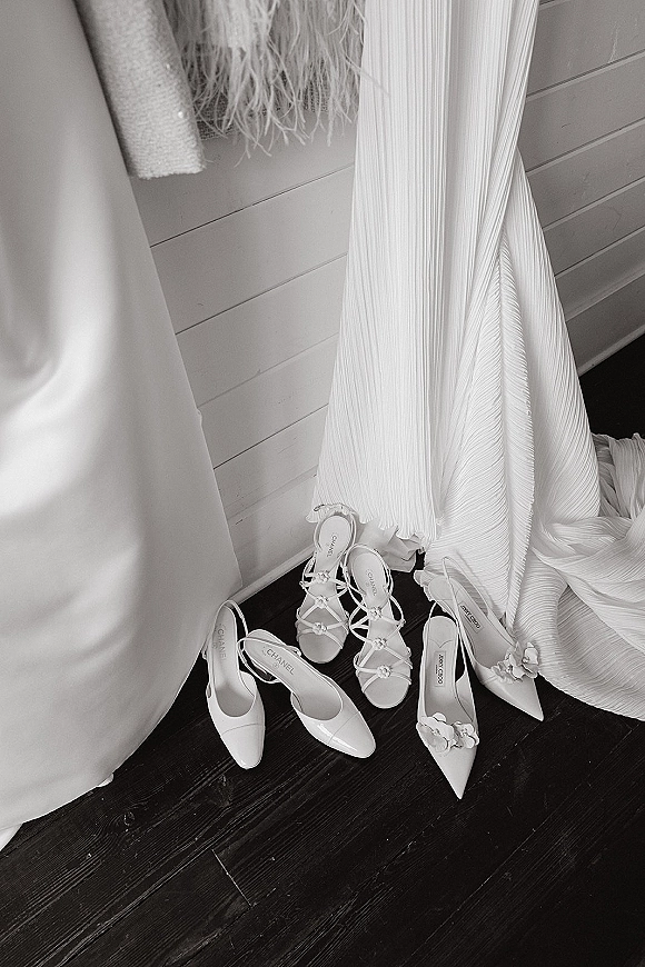 Wedding shoes flatlay with bridal shoes detail showing pointed toe heels and strappy sandals with floral appliques beside a pleated dress on wood floor