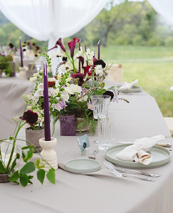 Reception tablescape with wedding table centerpiece, purple taper candles, and wax seal place cards on linen, set on a lawn with hills