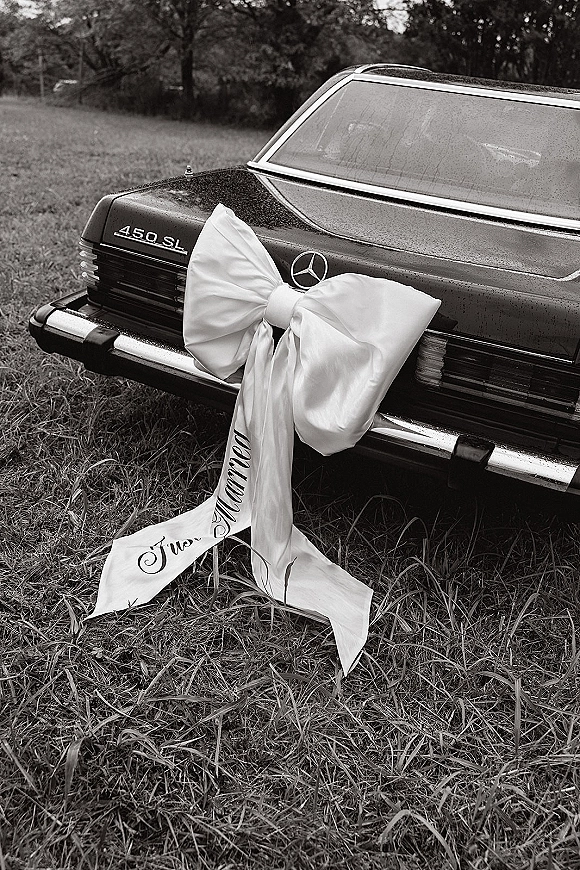 Wedding car decor with an oversized satin bow and ribbon streamers on a chrome bumper, parked by a grass field and trees