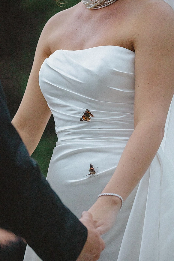 Wedding dress close-up of a strapless satin wedding dress with pearl necklace and veil, butterflies resting near the bodice outdoors