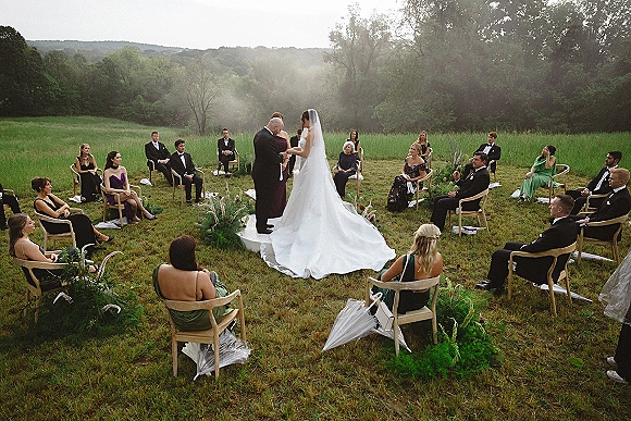 Wedding ceremony in a meadow with guests seated in a circle, bride in gown and veil with groom in tux, trees and hills beyond