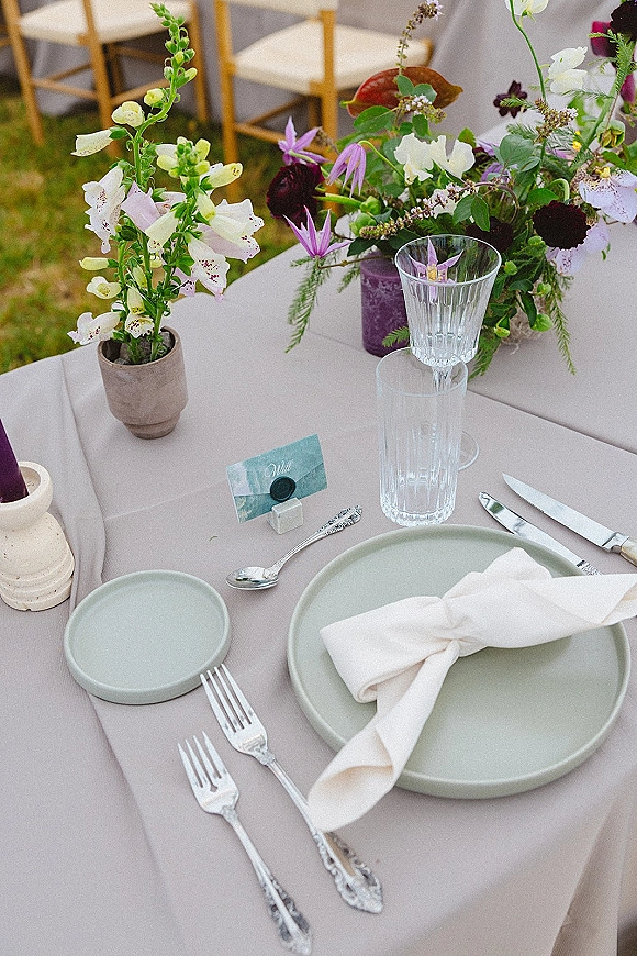 Reception tablescape with outdoor reception table place settings, purple taper candle and wildflower centerpiece on grass lawn under white drape