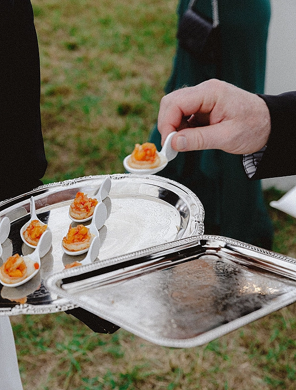 Wedding cocktail hour appetizers on a silver serving tray with ceramic tasting spoons, held by a server on an outdoor grass lawn