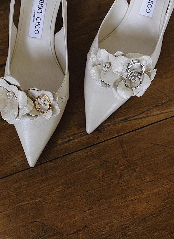 Wedding shoes detail with bridal shoes with rings, white pointed-toe heels and floral appliques on a wood floor flat lay