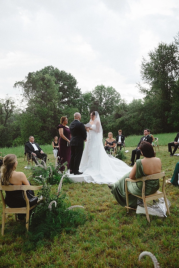 Wedding ceremony with couple exchanging vows as guests sit in a circle; bride’s cathedral veil and long train in a grassy field under overcast sky