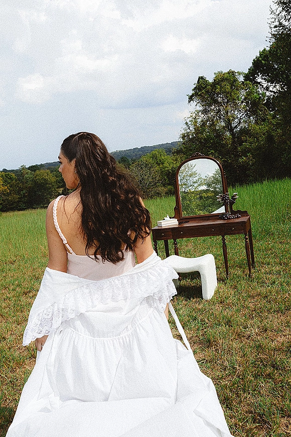 Bridal portrait of a bride from behind in an off the shoulder lace wedding dress and white knee-high boots, standing by a vanity in a grassy field