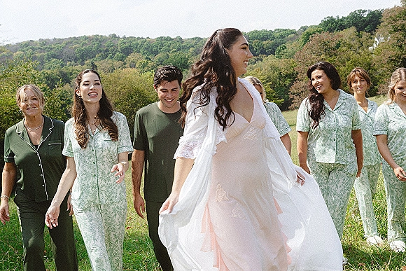 Bridal party portrait of bride with bridesmaids walking in green pajama sets, bride in white robe over dress, meadow hillside backdrop