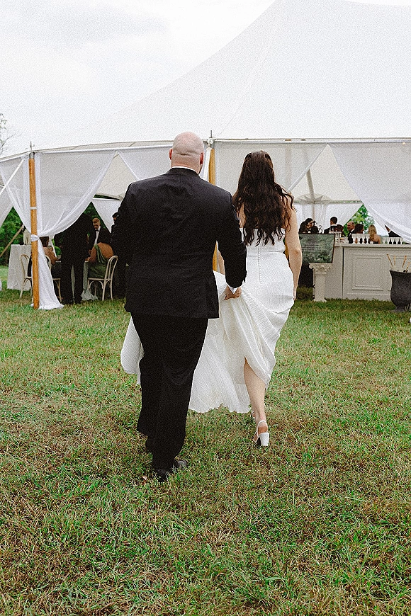 Wedding exit as bride and groom walk away holding hands, seen from behind under a clear-top draped tent with guests and bar setup