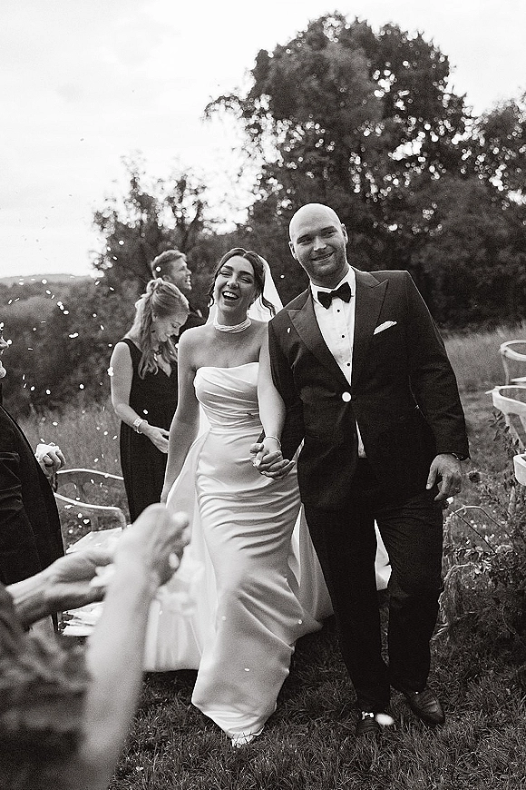 Wedding recessional as bride and groom walking hand in hand through confetti on an outdoor lawn aisle with guests and folding chairs