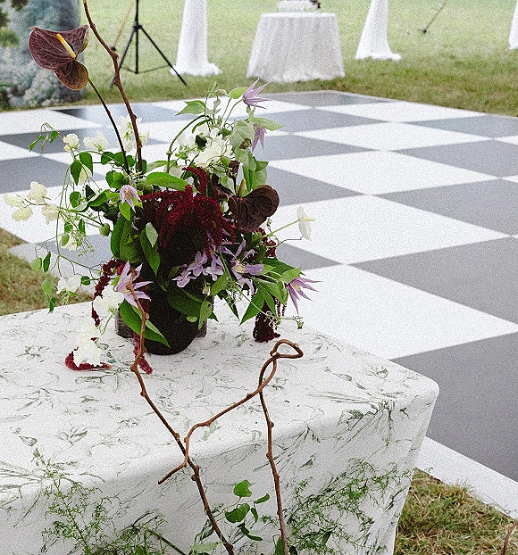 Wedding floral centerpiece in a black vase with burgundy, white, and purple blooms and greenery on a botanical print table by the lawn dance floor