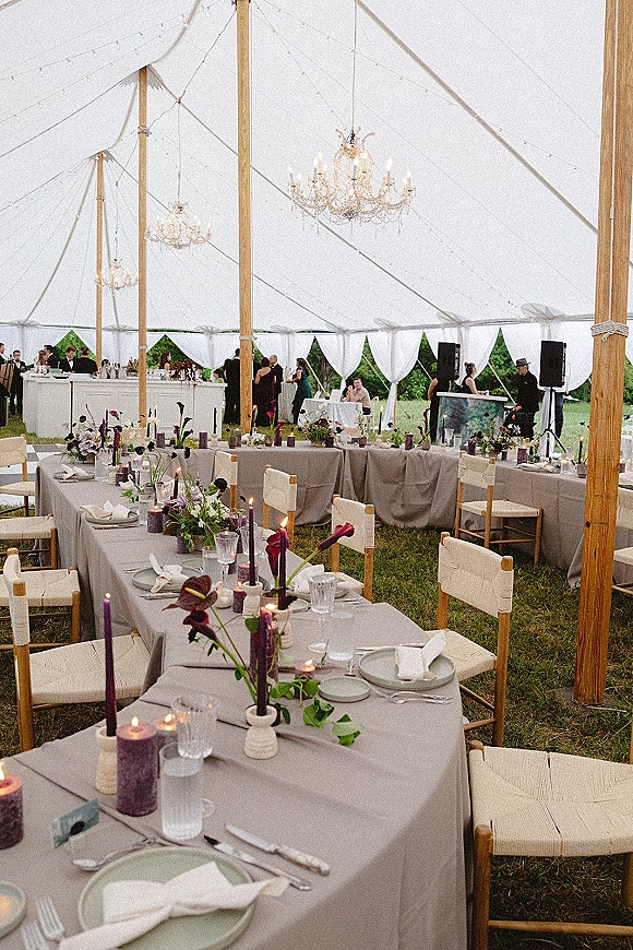 Reception tablescape with long banquet table setup, gray linens, candlelit floral centerpieces, woven chairs, and chandeliers under a draped tent