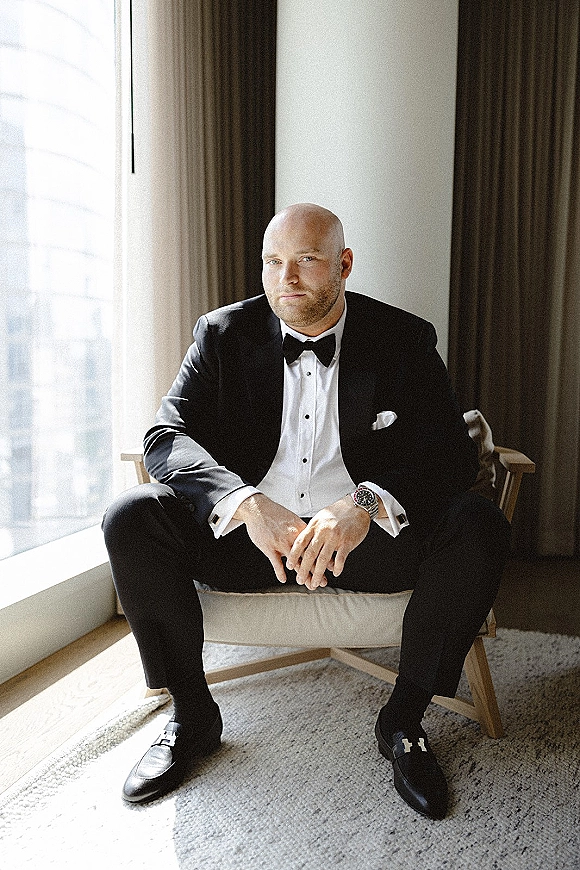 Groom portrait in a black tuxedo and bow tie, seated by window light in a hotel room, showing pocket square and wristwatch detail