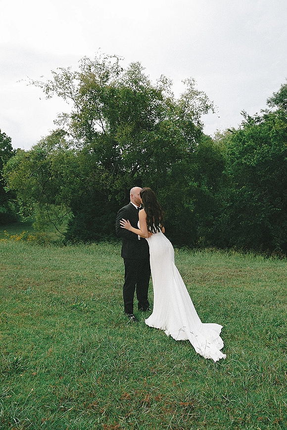 Wedding kiss portrait of bride and groom kissing as her veil and long dress train flow behind in a grassy field under overcast sky