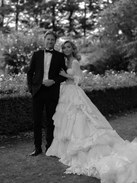 Couple portrait in a garden, bride in a long sleeve lace wedding dress with train, arm in arm with groom in tuxedo and bow tie
