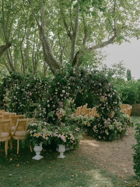Ceremony setup with outdoor wedding ceremony floral arch of garden roses and greenery, wooden chairs lining a gravel aisle under trees