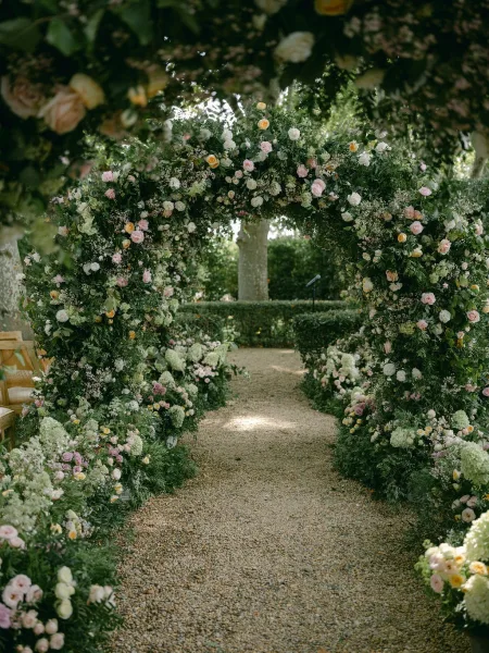 Ceremony arch decor with garden wedding floral arch of roses, hydrangeas, and greenery along a gravel aisle with wooden chairs and hedges