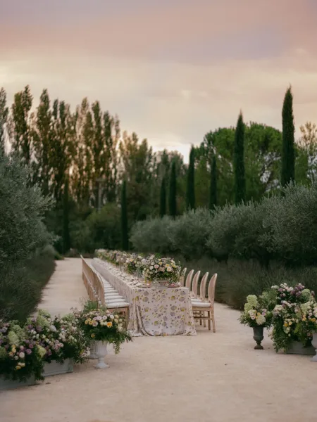 Reception tablescape with an outdoor banquet table lined with floral centerpieces and wooden chairs along a garden pathway under tall trees