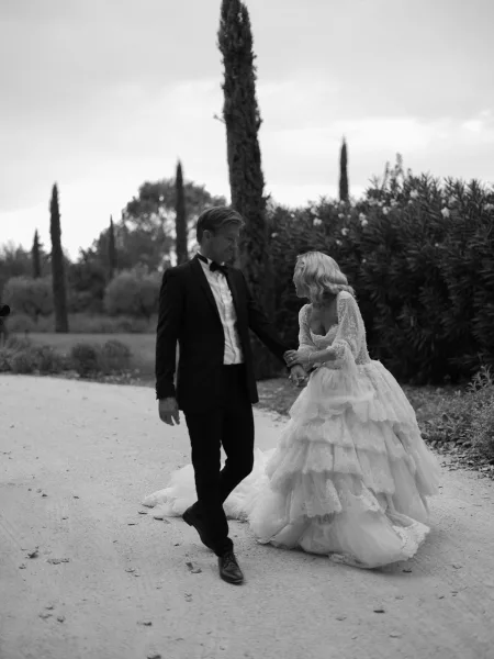 Couple portrait in black and white wedding portrait style, bride and groom walking hand in hand on a gravel path lined with tall trees