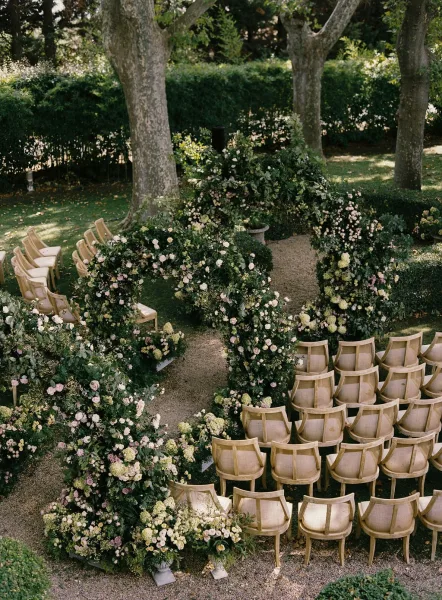 Ceremony setup with a floral arch and rose-lined gravel aisle, wooden chair rows on a garden lawn with trees and dappled sunlight