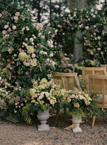 Ceremony floral decor featuring a wedding floral arch with roses, hydrangea and greenery, framed by urn planters and wooden chairs in a garden setting