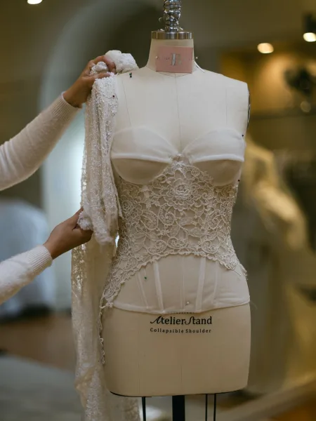 Wedding dress close-up of a bridal gown fitting on a mannequin as seamstress hands pin the lace corset bodice in a blurred atelier interior