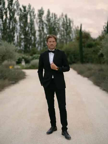 Groom portrait in a black tuxedo and bow tie, hands in pockets, standing on a garden path with trees under a cloudy sky
